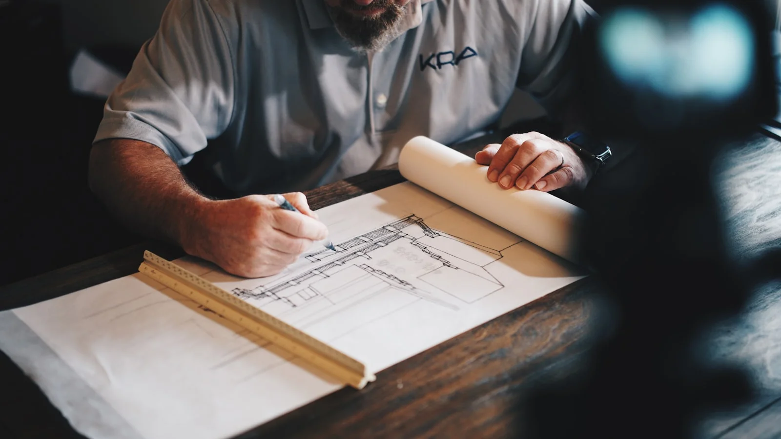 A solo contractor reviewing architectural building plans and a rolled blueprint at a warm wooden desk, calm and in control of the permit process.